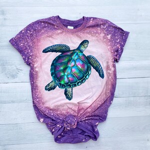 May include: A purple and pink tie-dye t-shirt with a bleached design featuring a colourful sea turtle.