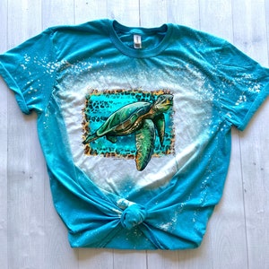 May include: A turquoise t-shirt with a bleached design and a sea turtle graphic. The turtle is green and brown, set against a turquoise and leopard print background. The shirt is knotted at the bottom.