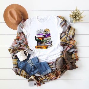 Peut inclure: T-shirt blanc avec un chat noir, un cr&acirc;ne et une pile de livres avec le mot "Spells" sur le livre du bas. Les livres sont d&eacute;cor&eacute;s d'une toile d'araign&eacute;e et d'autres d&eacute;tails sur le th&egrave;me d'Halloween.