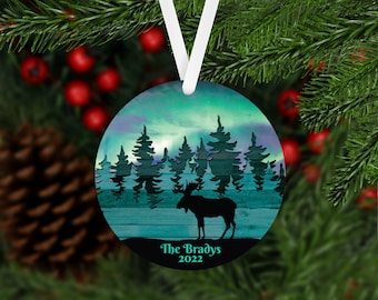 May include: Round Christmas ornament with a blue and green watercolor background featuring a silhouette of a moose in a forest. The text "The Bradys 2022" is printed at the bottom.