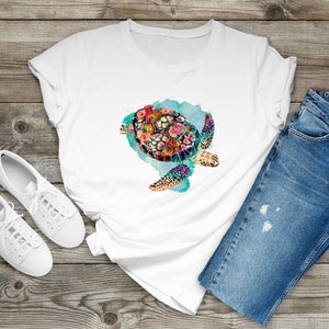Puede incluir: Camiseta blanca con un dise&ntilde;o de acuarela colorido de una tortuga marina con flores en su caparaz&oacute;n. La tortuga est&aacute; nadando en un fondo de acuarela azul y verde.