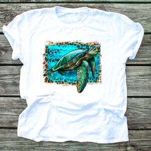 May include: White t-shirt featuring a detailed illustration of a sea turtle. The turtle is depicted in shades of green and brown, set against a turquoise background with a leopard print border. The shirt is made of a soft, comfortable material.