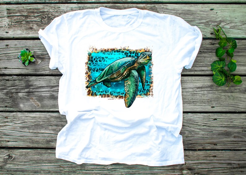 May include: White t-shirt featuring a detailed illustration of a sea turtle. The turtle is depicted in shades of green and brown, set against a turquoise background with a leopard print border. The shirt is made of a soft, comfortable material.