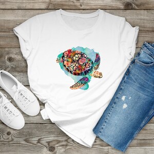 Puede incluir: Camiseta blanca con un dise&ntilde;o de acuarela colorido de una tortuga marina con flores en su caparaz&oacute;n. La tortuga est&aacute; nadando en un fondo de acuarela azul y verde.