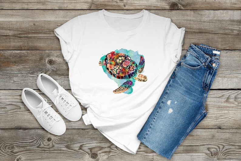 Puede incluir: Camiseta blanca con un dise&ntilde;o de acuarela colorido de una tortuga marina con flores en su caparaz&oacute;n. La tortuga est&aacute; nadando en un fondo de acuarela azul y verde.