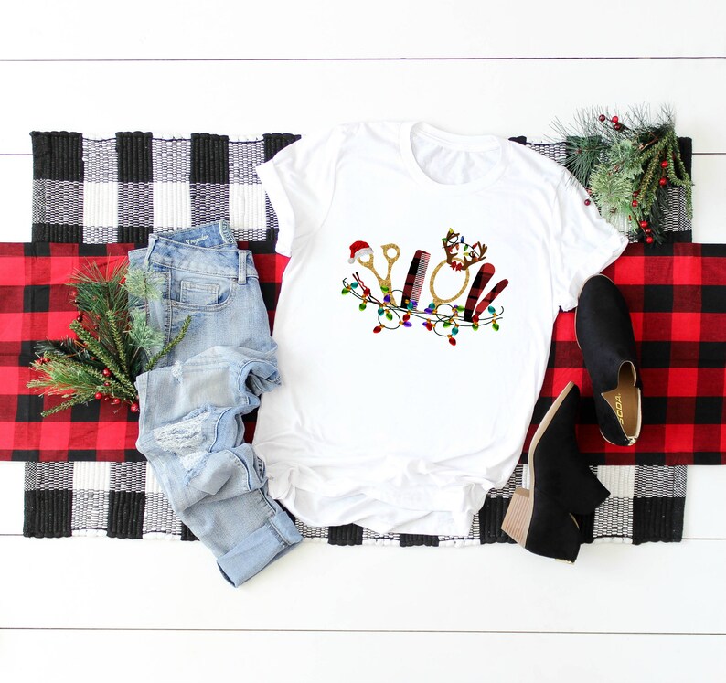 May include: White t-shirt with a Christmas-themed design featuring a pair of scissors, a hairbrush, a curling iron, and a comb. The design is surrounded by a garland of lights.