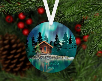 May include: Round Christmas ornament featuring a cabin in a forest with a reflection of the aurora borealis in a lake. The text "The Bradys 2022" is at the bottom of the ornament.