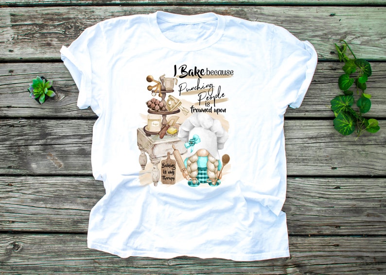 May include: White t-shirt with a baking-themed graphic. The design features a gnome chef, baking tools, and the text "I Bake because punching people is frowned upon. Baking is my therapy."