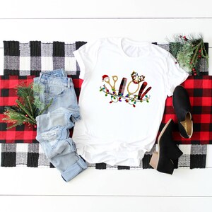May include: White t-shirt with a Christmas-themed design featuring a pair of scissors, a hairbrush, a curling iron, and a comb. The design is surrounded by a garland of lights.