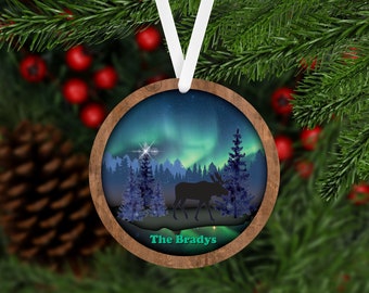 May include: A round wooden ornament with a silhouette of a moose standing in a forest with a green and blue aurora borealis in the background. The text "The Bradys" is written in green at the bottom of the ornament.