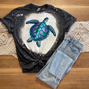 May include: A black and white tie-dye t-shirt featuring a colourful sea turtle graphic. The shirt has a distressed look with white bleach splatters.