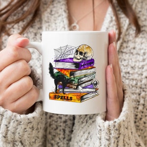 Peut inclure: Une tasse en c&eacute;ramique blanche avec un chat noir, un cr&acirc;ne et une pile de livres avec le mot "Spells" sur le livre du bas. Les livres sont d&eacute;cor&eacute;s d'une toile d'araign&eacute;e et d'autres d&eacute;tails sur le th&egrave;me d'Halloween.