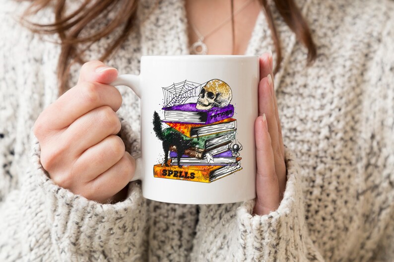 Peut inclure: Une tasse en c&eacute;ramique blanche avec un chat noir, un cr&acirc;ne et une pile de livres avec le mot "Spells" sur le livre du bas. Les livres sont d&eacute;cor&eacute;s d'une toile d'araign&eacute;e et d'autres d&eacute;tails sur le th&egrave;me d'Halloween.