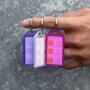 May include: Three keychains with different colored plastic frames and patterned paper inserts. The frames are purple, white, and pink. The paper inserts are a gradient of purple, a gradient of orange and pink, and a gradient of pink.