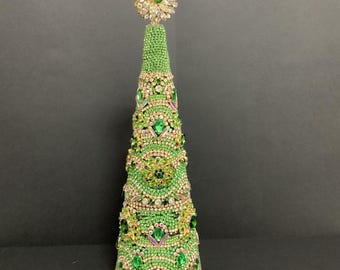 May include: A conical decorative tree sculpture covered in small green beads and various clear and green rhinestones. The surface features intricate patterns of embedded jewelry pieces, and the top is finished with a large, circular, star-shaped crystal brooch.