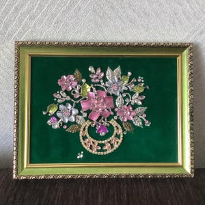May include: A framed floral artwork featuring a bouquet of pink, silver, and green flowers with rhinestone accents. The flowers are set against a dark green background within a gold-coloured frame.