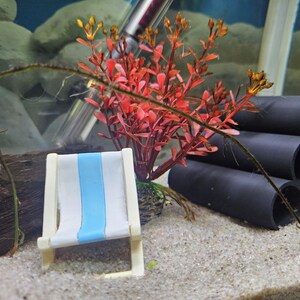 Könnte beinhalten: Ein weißer und blauer Miniatur-Strandstuhl mit einer roten und grünen Plastikpflanze in einem kleinen Topf. Der Stuhl steht auf einer weißen Sandfläche mit schwarzen Plastikrohren im Hintergrund.