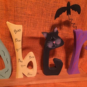 May include: Wooden Halloween decoration spelling out "Boo" with a teal, yellow, black, and purple color scheme. The letter "b" has "Boo" written on it multiple times. A black bat hangs above the "r".