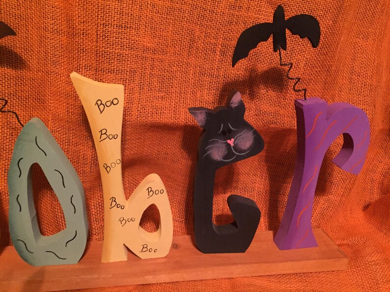 May include: Wooden Halloween decoration spelling out "Boo" with a teal, yellow, black, and purple color scheme. The letter "b" has "Boo" written on it multiple times. A black bat hangs above the "r".
