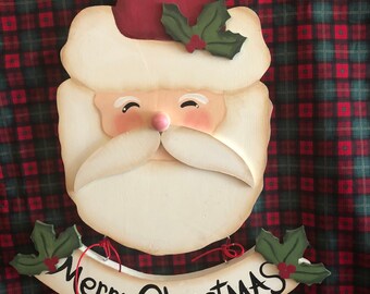 May include: Wooden Santa Claus Christmas decoration with a red hat, white beard, and a banner that reads "Merry Christmas". The decoration has holly accents and is set against a red and green plaid background.
