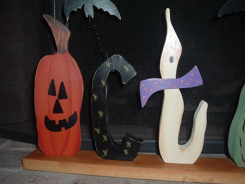 May include: Wooden Halloween decorations spelling out "OCT" with a pumpkin, a black letter "C" with gold stars, and a candle with a purple bow. The pumpkin is orange with a carved face. A black bat is above the letters.