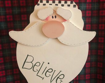 May include: Wooden Santa Claus face Christmas decoration. The Santa face is painted white with a red hat, pink nose, and white mustache. The word "Believe" is written in black script on the bottom of the face.