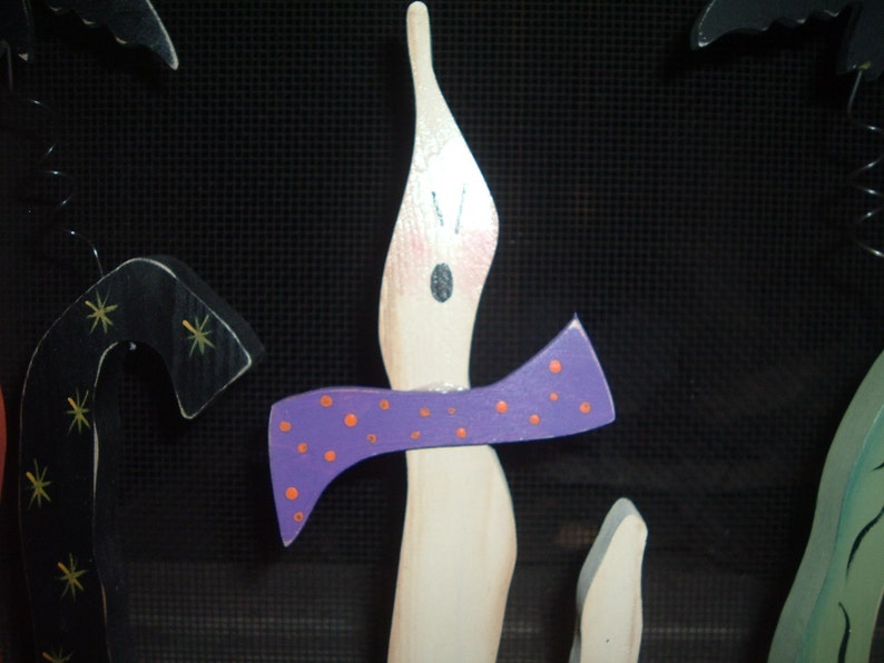May include: Wooden Halloween decorations featuring a candle with a purple bow and orange polka dots. Other decorations include a black bat and a black candy cane shape with gold stars. The background is black.