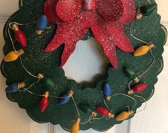 May include: A wooden Christmas wreath with a dark green base and a large red bow. The wreath is decorated with colorful lights in red, yellow, blue, and green. The bow has white speckles, and the lights are strung on gold wire.