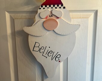 May include: White wooden Santa Claus head with a red hat, black and white trim, and a white beard. The word "Believe" is written in black on the beard with a small red heart.