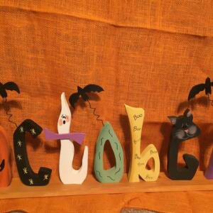 May include: Wooden Halloween decoration spelling "October" with a pumpkin, black cat, ghost, and bats. The letters are painted in orange, black, white, green, yellow, and purple. The word "Boo" is written on the letter "b".