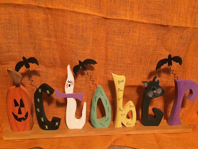 May include: Wooden Halloween decoration spelling "October" with a pumpkin, black cat, ghost, and bats. The letters are painted in orange, black, white, green, yellow, and purple. The word "Boo" is written on the letter "b".