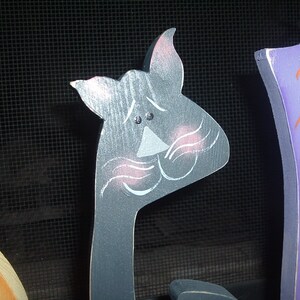 May include: A gray wooden cat head with pink blush marks and white whiskers. The cat is part of a Halloween decoration. The cat's eyes are small black dots. The cat is painted on a wooden cutout.
