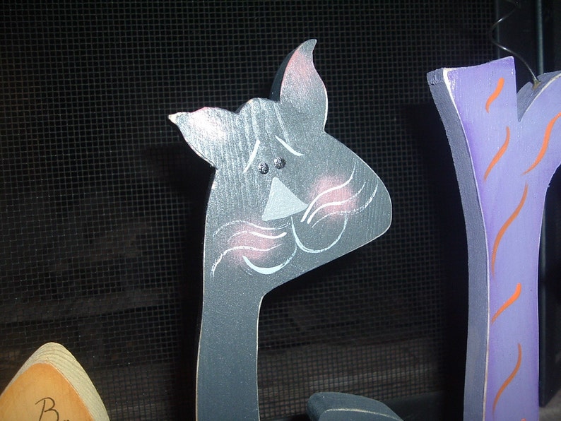 May include: A gray wooden cat head with pink blush marks and white whiskers. The cat is part of a Halloween decoration. The cat's eyes are small black dots. The cat is painted on a wooden cutout.