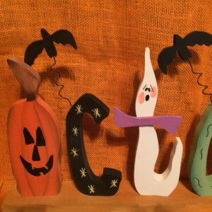 May include: Wooden Halloween-themed decoration spelling out the word "BOO". The orange pumpkin, black "C", white ghost, and green teardrop shapes are topped with black bats. The background is orange.