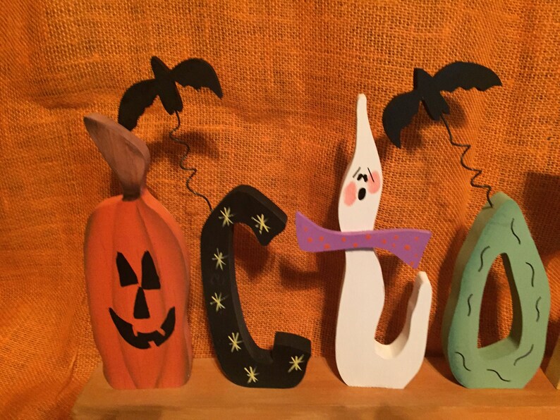 May include: Wooden Halloween-themed decoration spelling out the word "BOO". The orange pumpkin, black "C", white ghost, and green teardrop shapes are topped with black bats. The background is orange.