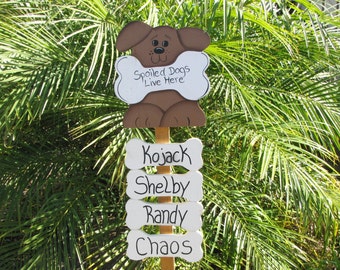 May include: A wooden sign shaped like a dog with a bone in its mouth. The sign reads "Spoiled Dogs Live Here". Below the dog are four white bone-shaped signs with the names "Kojack", "Shelby", "Randy", and "Chaos" written in black.