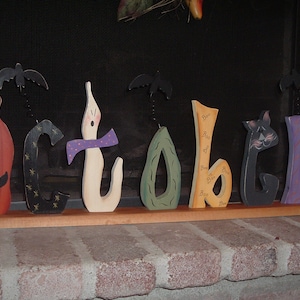 May include: Wooden Halloween decoration spelling "October". The letters are painted in various colors and feature a jack-o'-lantern, ghost, and cat. Each letter has a unique design, with bats attached above some letters.