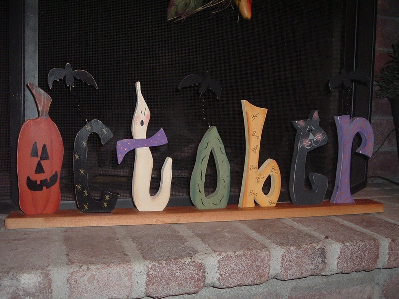 May include: Wooden Halloween decoration spelling "October". The letters are painted in various colors and feature a jack-o'-lantern, ghost, and cat. Each letter has a unique design, with bats attached above some letters.