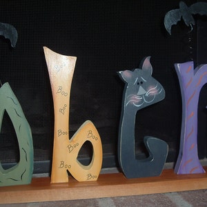 May include: Wooden Halloween decoration spelling out "Boo" with a green, orange, black, and purple color scheme. The orange letter has "Boo" written on it multiple times. Two black bats hang above the letters.