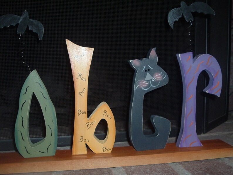 May include: Wooden Halloween decoration spelling out "Boo" with a green, orange, black, and purple color scheme. The orange letter has "Boo" written on it multiple times. Two black bats hang above the letters.