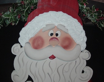 May include: A wooden Santa Claus face decoration with a red hat and white beard. The face has rosy cheeks, a red mouth, and black eyes. The beard is a swirling design. Red ornaments hang below.