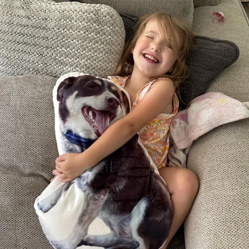 May include: A smiling child hugs a custom pillow featuring a dog's portrait. The pillow showcases the dog with a brown and white coat, blue collar, and open mouth. The child is wearing a floral print top.