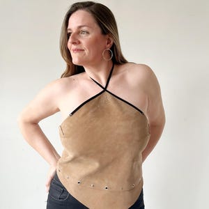 May include: A tan suede halter top featuring a pointed hemline, thin black straps, and silver-toned metal grommets along the lower edge. The garment has a triangular front panel with a small cluster of silver studs near the neckline.