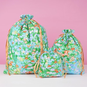 May include: Three drawstring bags in varying sizes. The bags are covered in a floral pattern with green, blue, and orange flowers on a light blue background. Each bag has a gold-colored drawstring closure.
