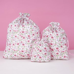 Può includere: Tre sacchetti con coulisse di diverse dimensioni, in tessuto di cotone, con motivo floreale rosa e bianco. I sacchetti hanno un cordoncino rosa.