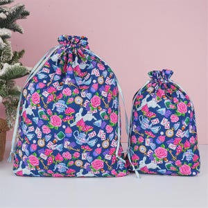 May include: Two drawstring bags with an Alice in Wonderland design. The bags have a navy blue background with pink roses, white rabbits, teacups, and playing cards. The bags have light blue drawstrings.