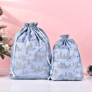 May include: Two blue drawstring bags with a silver and white winter scene print. The larger bag has a light blue drawstring and the smaller bag has a white drawstring.
