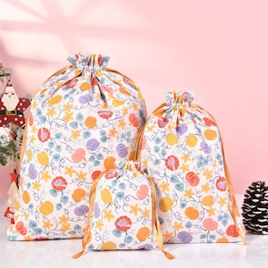 May include: Three white drawstring bags with a pumpkin and vine pattern. The bags are different sizes and have gold drawstrings.