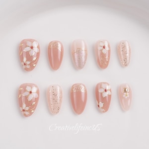 May include: A set of ten artificial nails in various shades of pink and nude. Some nails feature white floral designs, gold accents, or glitter. The nails are oval-shaped and arranged on a white surface.