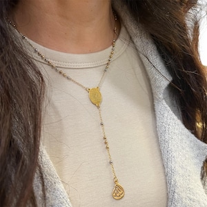 May include: Gold-coloured necklace with a drop pendant. The necklace features small beads and a central oval-shaped pendant. The drop pendant has a teardrop shape with an engraved design. The necklace is worn on a person.